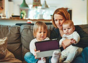 two babies and woman sitting on sofa while holding baby and watching on tablet