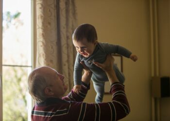 man wearing maroon, white, and blue stripe long-sleeved shirt lifting up baby wearing gray onesie