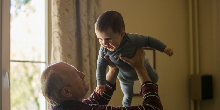 man wearing maroon, white, and blue stripe long-sleeved shirt lifting up baby wearing gray onesie